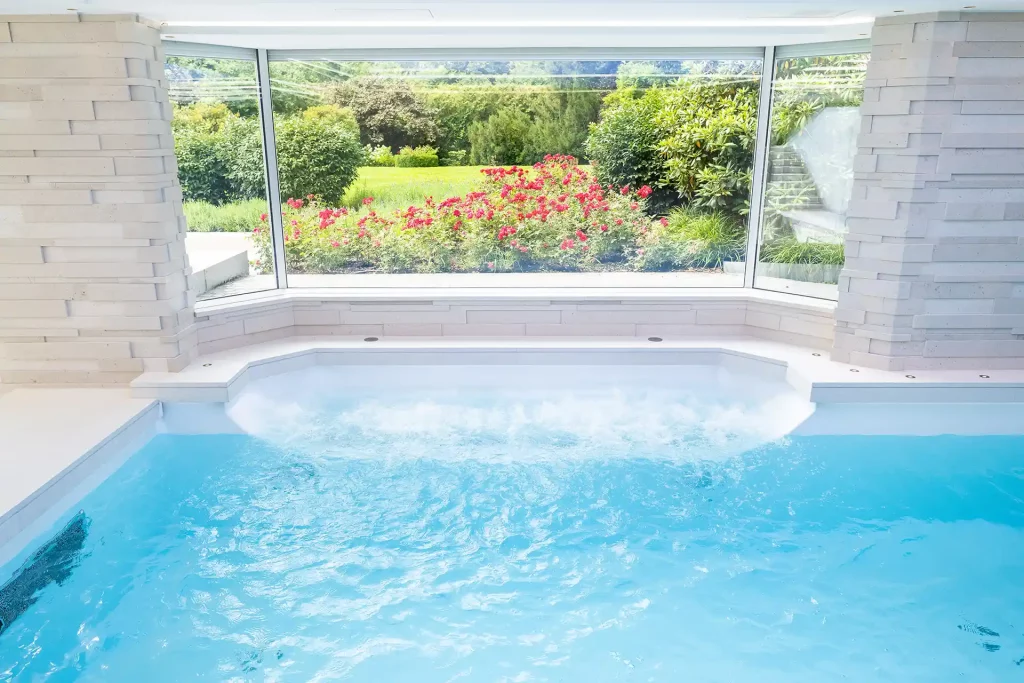 Ein moderner Indoor-Swimmingpool mit integrierter Massagestation und einem großen Panoramafenster mit Blick in den Garten.